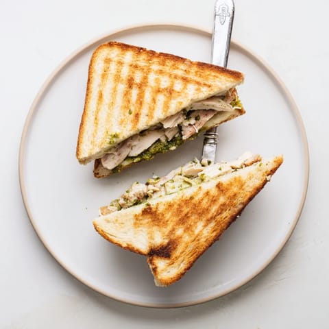 The Mozzarella Pesto Chicken Grilled Cheese is cut in half on a wooden board, revealing layers of shredded chicken, pesto, and fresh mozzarella.  