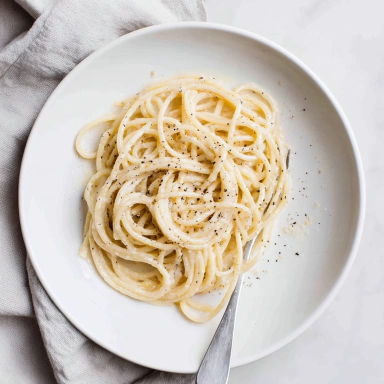 A white bowl holds a serving of Spaghetti Cacio e Pepe, garnished with extra Pecorino and a lemon wedge.