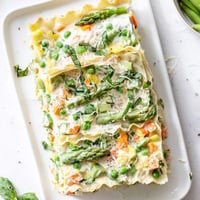 Golden-brown Spring Veggie Alfredo Lasagna baked bubbly in a dish, layered with creamy sauce, tender asparagus, carrots, and fresh basil garnish.